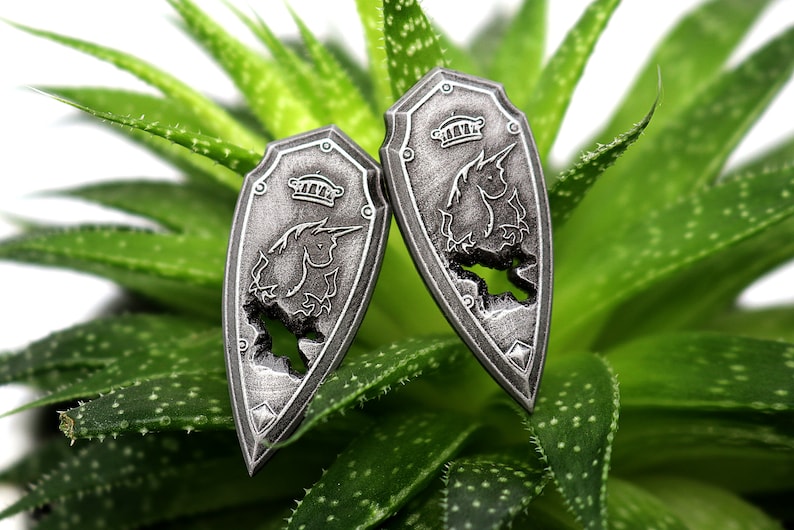 May include: Two silver-colored shield-shaped earrings with a unicorn design. The unicorn is outlined in the shield and has a crown on its head.