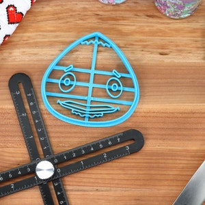 May include: A blue plastic cookie cutter shaped like a cartoon face with a wide smile and large eyes. The cutter is on a wooden surface.