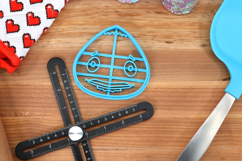 May include: A blue plastic cookie cutter shaped like a cartoon face with a wide smile and large eyes. The cutter is on a wooden surface.