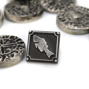 May include: A silver metal token with a stylized fish design on a black background. The token is surrounded by other silver metal tokens with intricate designs.