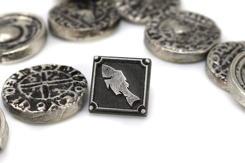 May include: A silver metal token with a stylized fish design on a black background. The token is surrounded by other silver metal tokens with intricate designs.