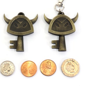 May include: Two gold-colored keychains shaped like a horned helmet with a key attached. The keychains are on a white surface with three coins next to them.