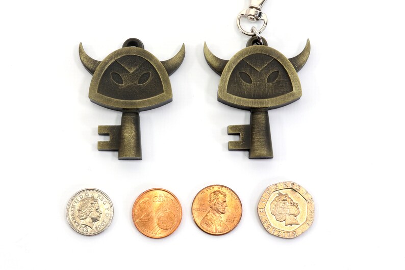 May include: Two gold-colored keychains shaped like a horned helmet with a key attached. The keychains are on a white surface with three coins next to them.