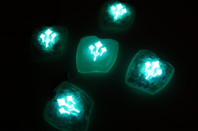 May include: Five teal green resin heart-shaped ornaments with a glowing white floral design. The ornaments are on a black background.