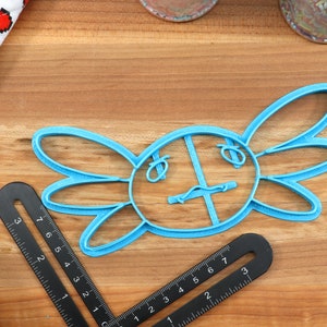 May include: Blue plastic cookie cutter shaped like an axolotl, a type of salamander. The cutter has a round body with two large, rounded fins on either side. The axolotl has a small mouth and two small eyes.