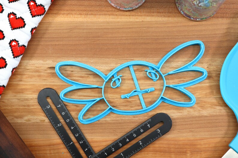 May include: Blue plastic cookie cutter shaped like an axolotl, a type of salamander. The cutter has a round body with two large, rounded fins on either side. The axolotl has a small mouth and two small eyes.