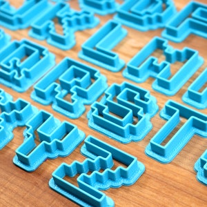 May include: A set of blue plastic cookie cutters shaped like letters of the alphabet in a pixelated style.