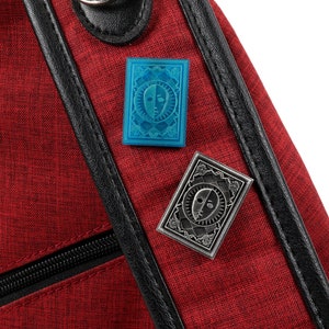 Op de afbeelding: Twee emaillepins met een gestileerd gezicht met twee kanten, een lichte en een donkere. De pins zijn bevestigd aan een rode stoffen tas met een zwarte rits en zwarte bies.