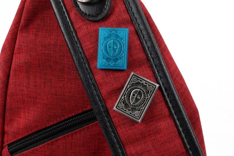 Op de afbeelding: Twee emaillepins met een gestileerd gezicht met twee kanten, een lichte en een donkere. De pins zijn bevestigd aan een rode stoffen tas met een zwarte rits en zwarte bies.