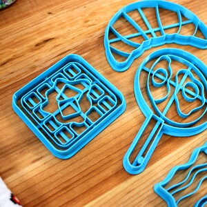 May include: A collection of bright blue cookie cutters in various shapes, including a square, a frying pan, and a wavy design, are arranged on a wooden surface. The cutters are made of plastic and are designed for baking. Sprinkles are visible in the background.
