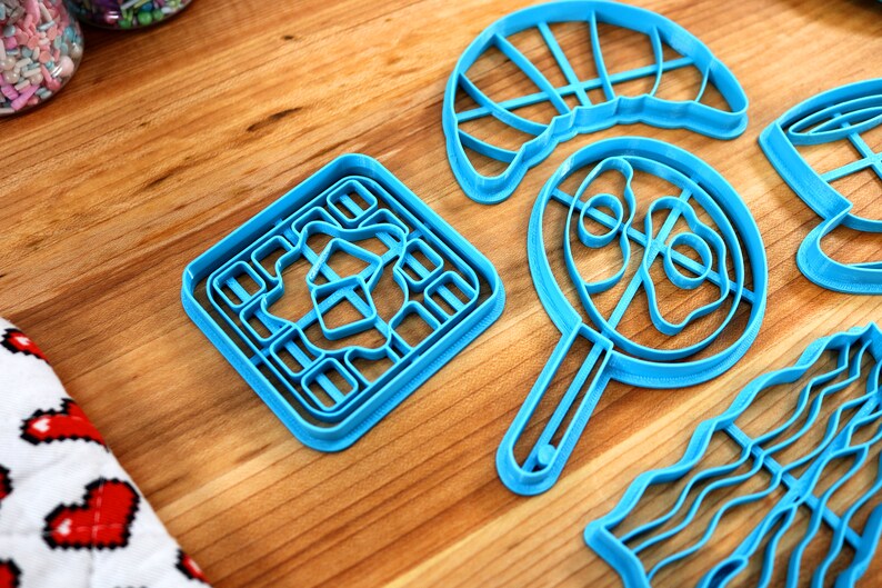May include: A collection of bright blue cookie cutters in various shapes, including a square, a frying pan, and a wavy design, are arranged on a wooden surface. The cutters are made of plastic and are designed for baking. Sprinkles are visible in the background.