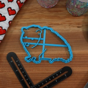 May include: A blue plastic cookie cutter shaped like a hamster. The cutter is on a wooden surface.