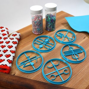 May include: A set of six blue cookie cutters with different designs, a red oven mitt, a rolling pin, two jars of sprinkles, and a blue spatula on a wooden cutting board.