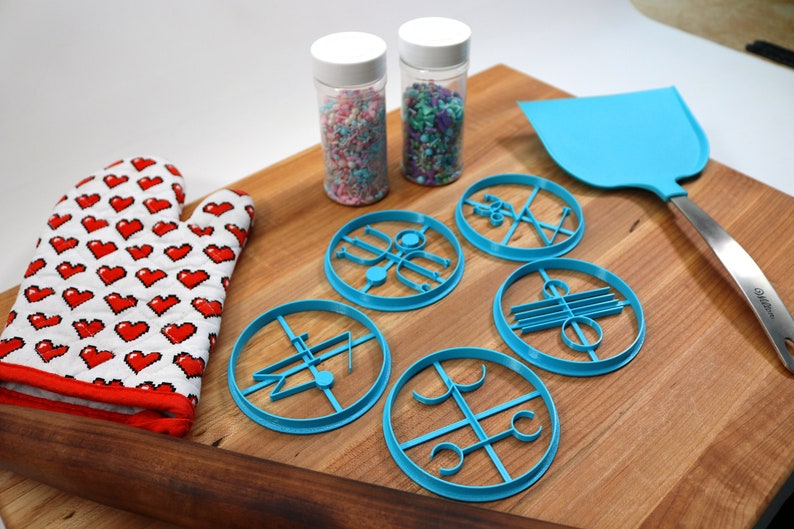 May include: A set of six blue cookie cutters with different designs, a red oven mitt, a rolling pin, two jars of sprinkles, and a blue spatula on a wooden cutting board.