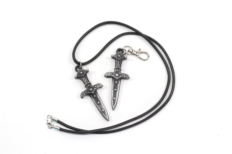 May include: Two silver-toned dagger pendants on a black cord necklace. The necklace has a silver clasp.
