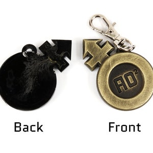 May include: A black and gold metal keychain with a round charm featuring the text "AD&sup2;" in black. The back of the charm has a stylized design.