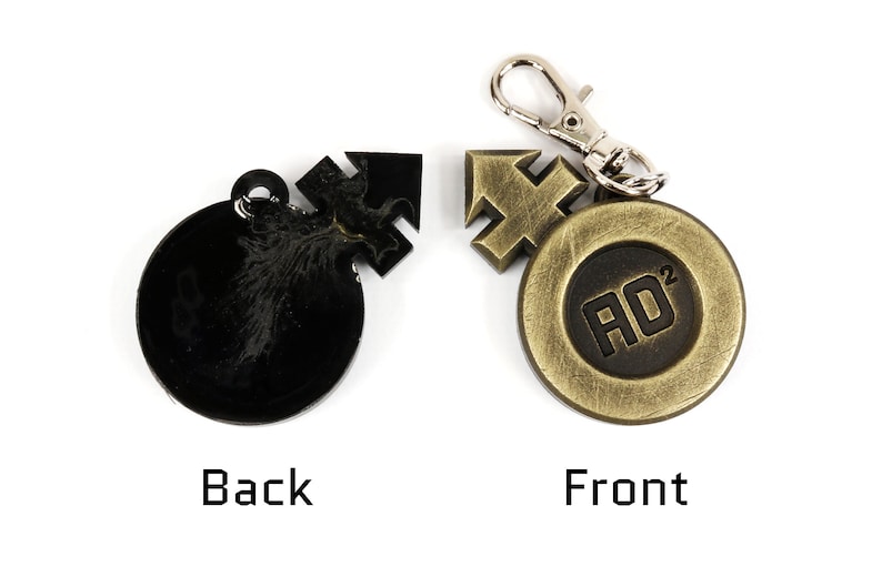 May include: A black and gold metal keychain with a round charm featuring the text "AD&sup2;" in black. The back of the charm has a stylized design.