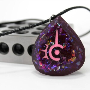 May include: A teardrop-shaped pendant necklace with a black cord. The pendant is a deep purple with iridescent glitter and a pink symbol. The necklace is on a white surface with a silver metal object in the background.