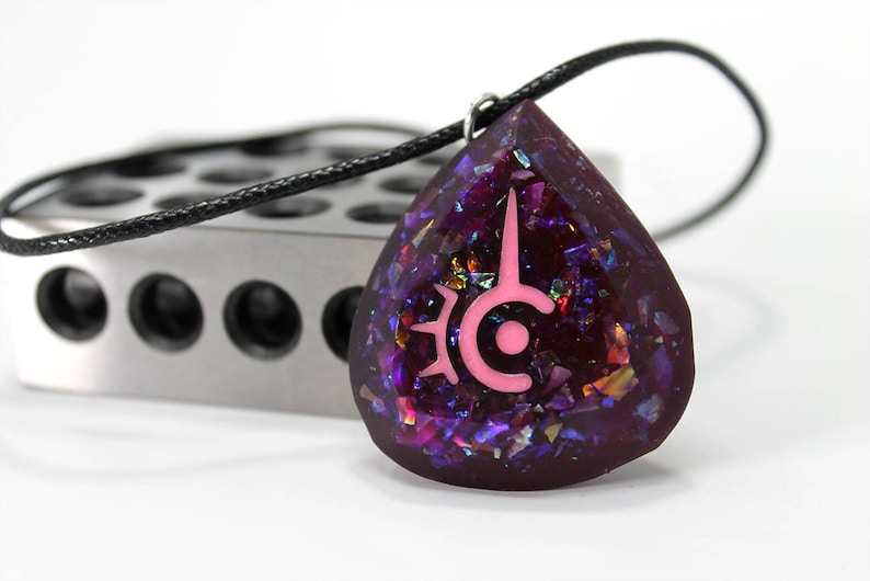May include: A teardrop-shaped pendant necklace with a black cord. The pendant is a deep purple with iridescent glitter and a pink symbol. The necklace is on a white surface with a silver metal object in the background.
