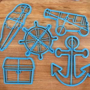 May include: A collection of blue plastic cookie cutters in nautical shapes, including a parrot, ship's wheel, cannon, anchor, and treasure chest. The cutters are arranged on a wooden surface, suggesting a pirate or seafaring theme. A blue spatula is visible.