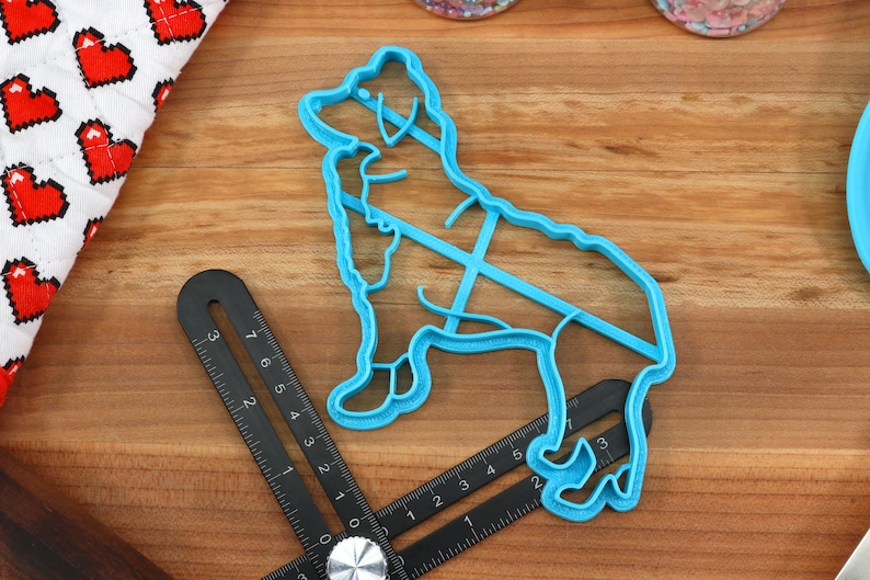 May include: A blue plastic cookie cutter shaped like a dog. The cutter is on a wooden surface with a black ruler.