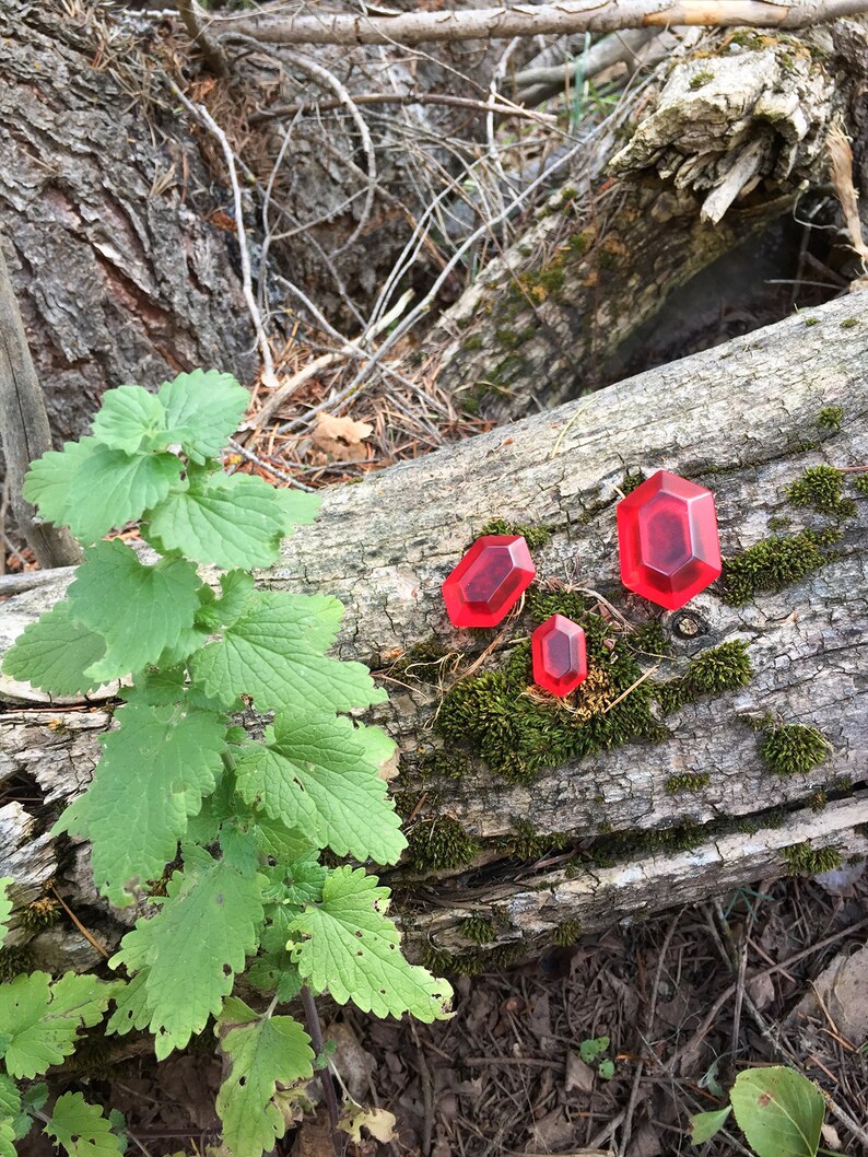 May include: Three red plastic gems, shaped like a rhombus, are arranged on a fallen log in a forest setting. The gems are different sizes, with the largest gem in the center and the smallest gem on the left. The log is covered in moss and surrounded by green leaves.