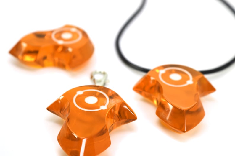 May include: Three orange glass pendants with a white atom symbol design. Two pendants are on a white surface, and one is on a black cord.