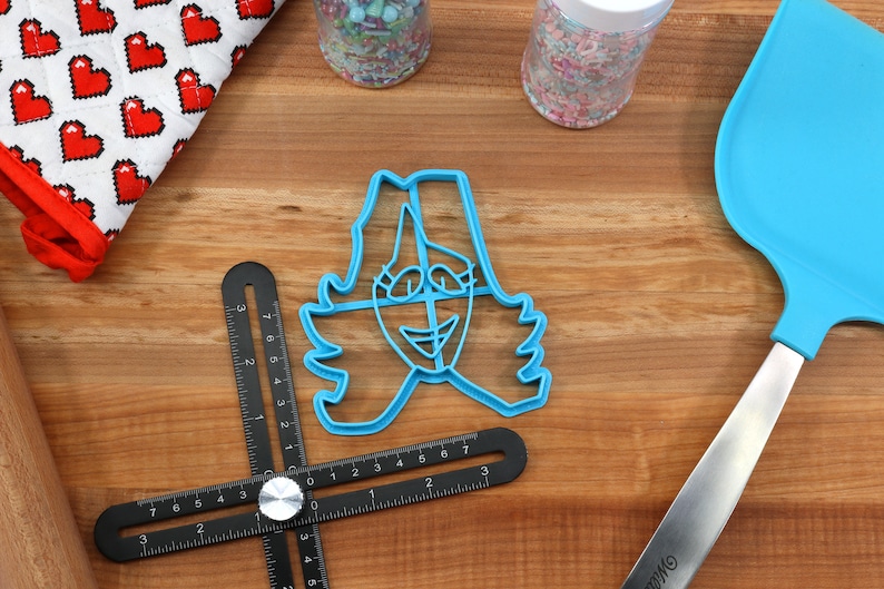 May include: Blue plastic cookie cutter shaped like a person with a smiling face and wavy hair. The cutter is on a wooden surface with a ruler and a spatula.