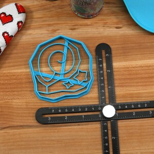 May include: Blue plastic cookie cutter shaped like a crescent moon with stars. The cutter is on a wooden surface with a black ruler.