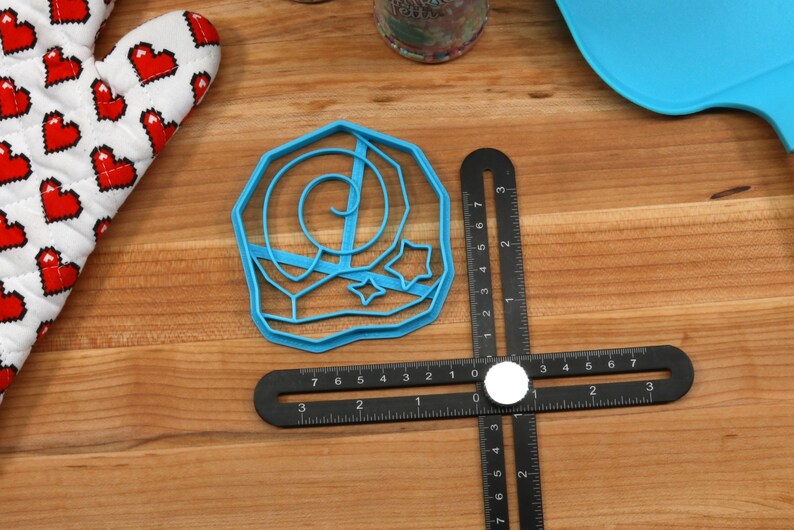 May include: Blue plastic cookie cutter shaped like a crescent moon with stars. The cutter is on a wooden surface with a black ruler.