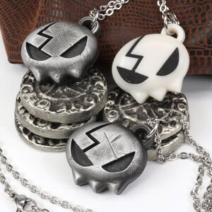 May include: Two silver skull necklaces with a cracked design. One skull is white and the other is silver. The necklaces are on a brown leather background.
