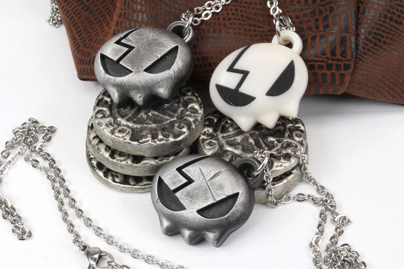 May include: Two silver skull necklaces with a cracked design. One skull is white and the other is silver. The necklaces are on a brown leather background.