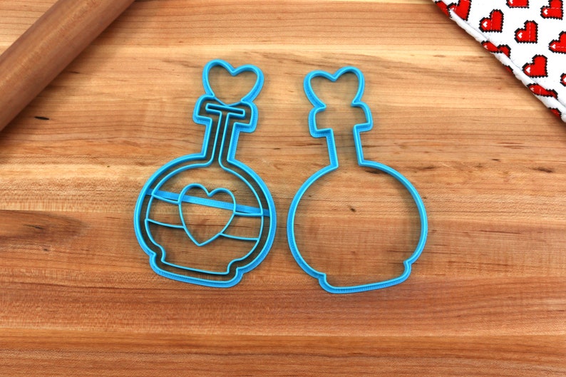 May include: Two blue plastic cookie cutters shaped like potion bottles with heart details. One cutter has a heart design on the bottle and the other is plain.