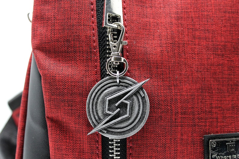 May include: A silver metal zipper charm with a stylized lightning bolt and a circular design. The charm is attached to a red backpack.