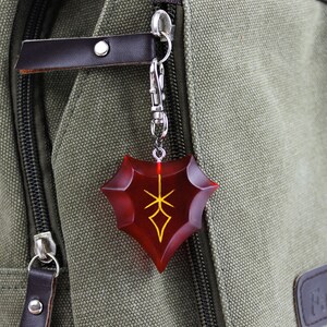 May include: A red and gold shield-shaped keychain with a stylized design. The keychain is attached to a silver chain and is hanging from a green canvas bag.
