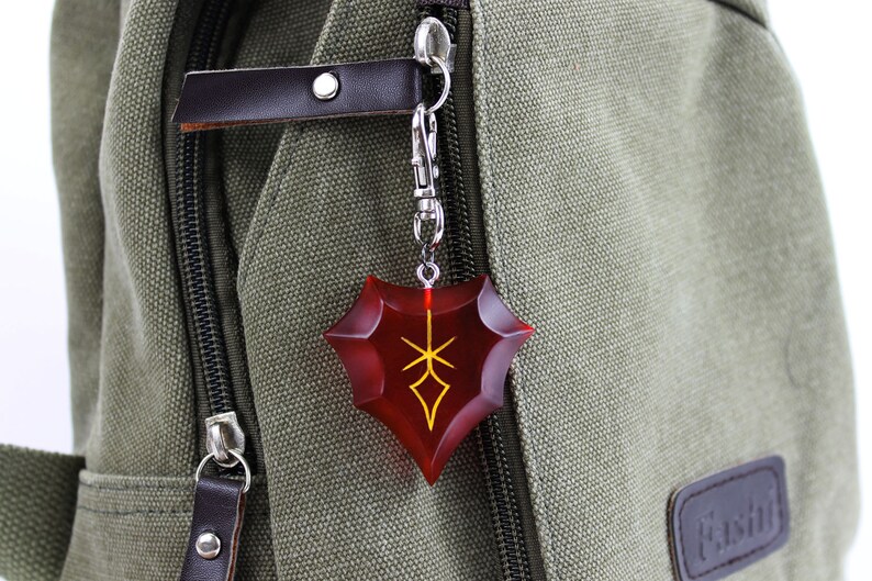 May include: A red and gold shield-shaped keychain with a stylized design. The keychain is attached to a silver chain and is hanging from a green canvas bag.