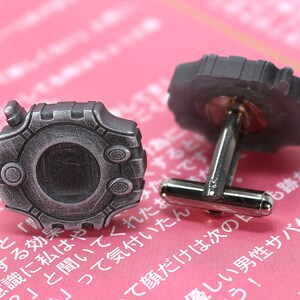 May include: A pair of silver cufflinks with a circular design. The cufflinks feature a detailed, textured surface with a raised center and a small, circular opening. The cufflinks are on a pink background.