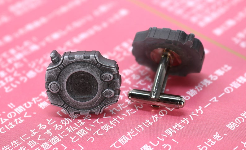 May include: A pair of silver cufflinks with a circular design. The cufflinks feature a detailed, textured surface with a raised center and a small, circular opening. The cufflinks are on a pink background.