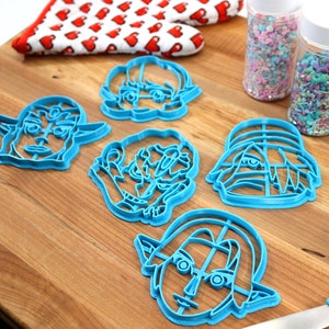 May include: Five blue plastic cookie cutters shaped like cartoon faces on a wooden surface. The cutters are for making cookies in the shape of faces.