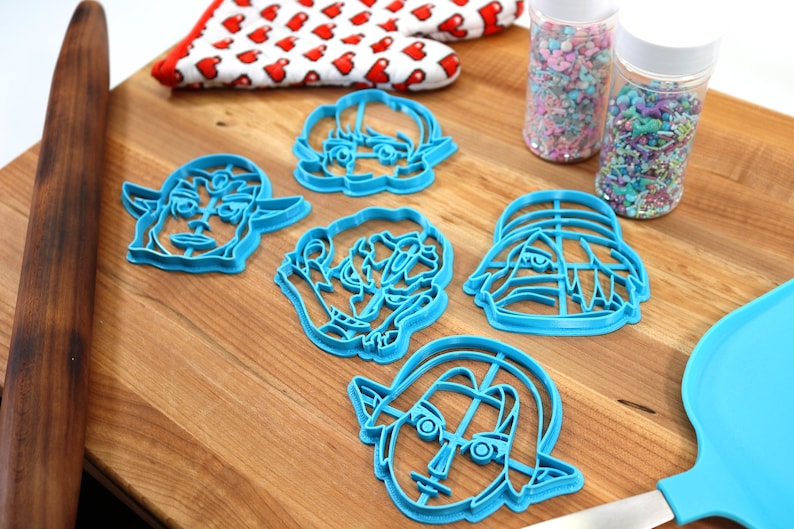 May include: Five blue plastic cookie cutters shaped like cartoon faces on a wooden surface. The cutters are for making cookies in the shape of faces.
