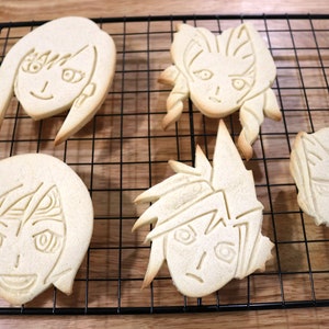 May include: Five decorated sugar cookies, each featuring a different anime character, are arranged on a black wire cooling rack. The cookies are light brown and have a slightly glossy finish.