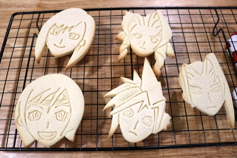 May include: Five decorated sugar cookies, each featuring a different anime character, are arranged on a black wire cooling rack. The cookies are light brown and have a slightly glossy finish.