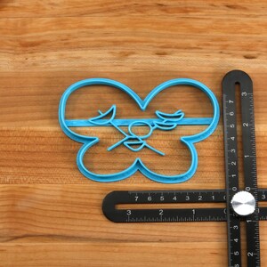 May include: A blue plastic cookie cutter shaped like a butterfly with a stylized design in the center. The cutter is on a wooden surface with a black ruler.