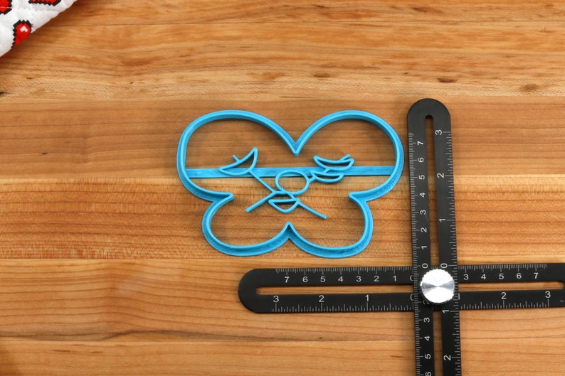 May include: A blue plastic cookie cutter shaped like a butterfly with a stylized design in the center. The cutter is on a wooden surface with a black ruler.