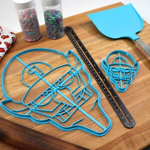 May include: A set of blue plastic cookie cutters shaped like a face and a smaller face with a hat. The cutters are on a wooden cutting board with a rolling pin, a spatula, two jars of sprinkles, and a red and white oven mitt.