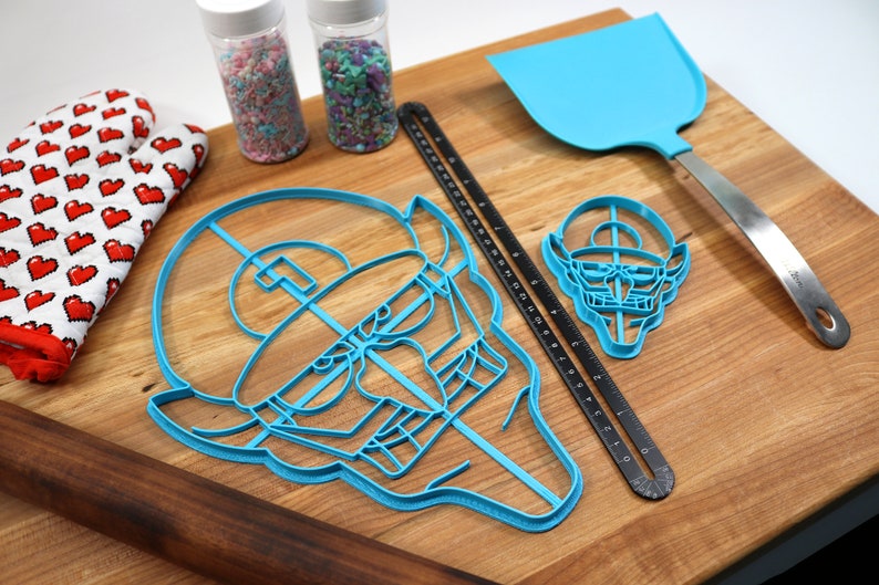 May include: A set of blue plastic cookie cutters shaped like a face and a smaller face with a hat. The cutters are on a wooden cutting board with a rolling pin, a spatula, two jars of sprinkles, and a red and white oven mitt.