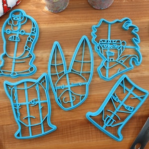 May include: Set of six blue plastic cookie cutters shaped like cartoon characters. The cutters are on a wooden surface.