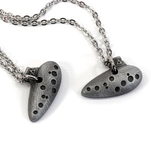 May include: Two silver ocarina necklaces with silver chains. The ocarinas are in the shape of a heart and a teardrop.