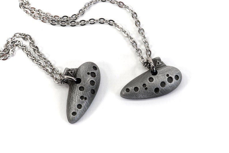 May include: Two silver ocarina necklaces with silver chains. The ocarinas are in the shape of a heart and a teardrop.