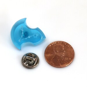 May include: A blue resin pin with a white outline of the constellation Pisces. The pin is about the size of a US penny.